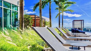 Un área de lounge en la azotea con tumbonas y mesas, palmeras y un edificio moderno con grandes ventanales.