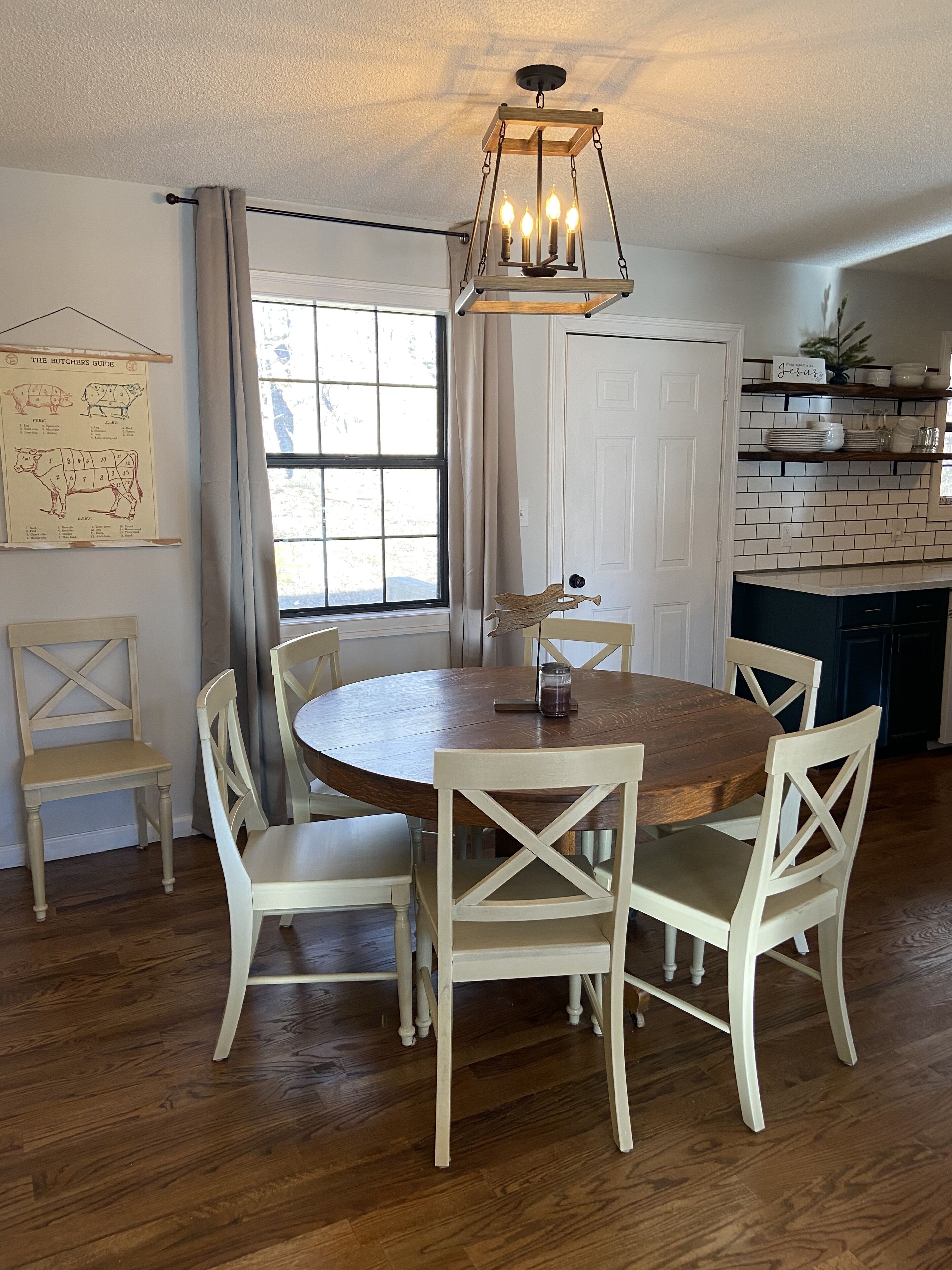 Un coin repas avec une table ronde en bois et des chaises blanches, un luminaire suspendu, un tableau explicatif sur la découpe de la viande accroché au mur, et une fenêtre avec des rideaux.