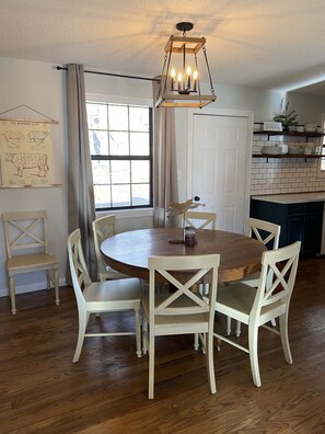 A dining area with a round wooden table and white chairs, a hanging light fixture, a butcher's guide on the wall, and a window with curtains.