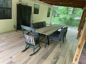 A covered outdoor patio with a wooden table and chairs, a rocking chair with a plaid cushion, a black cabinet, and a window overlooking greenery.