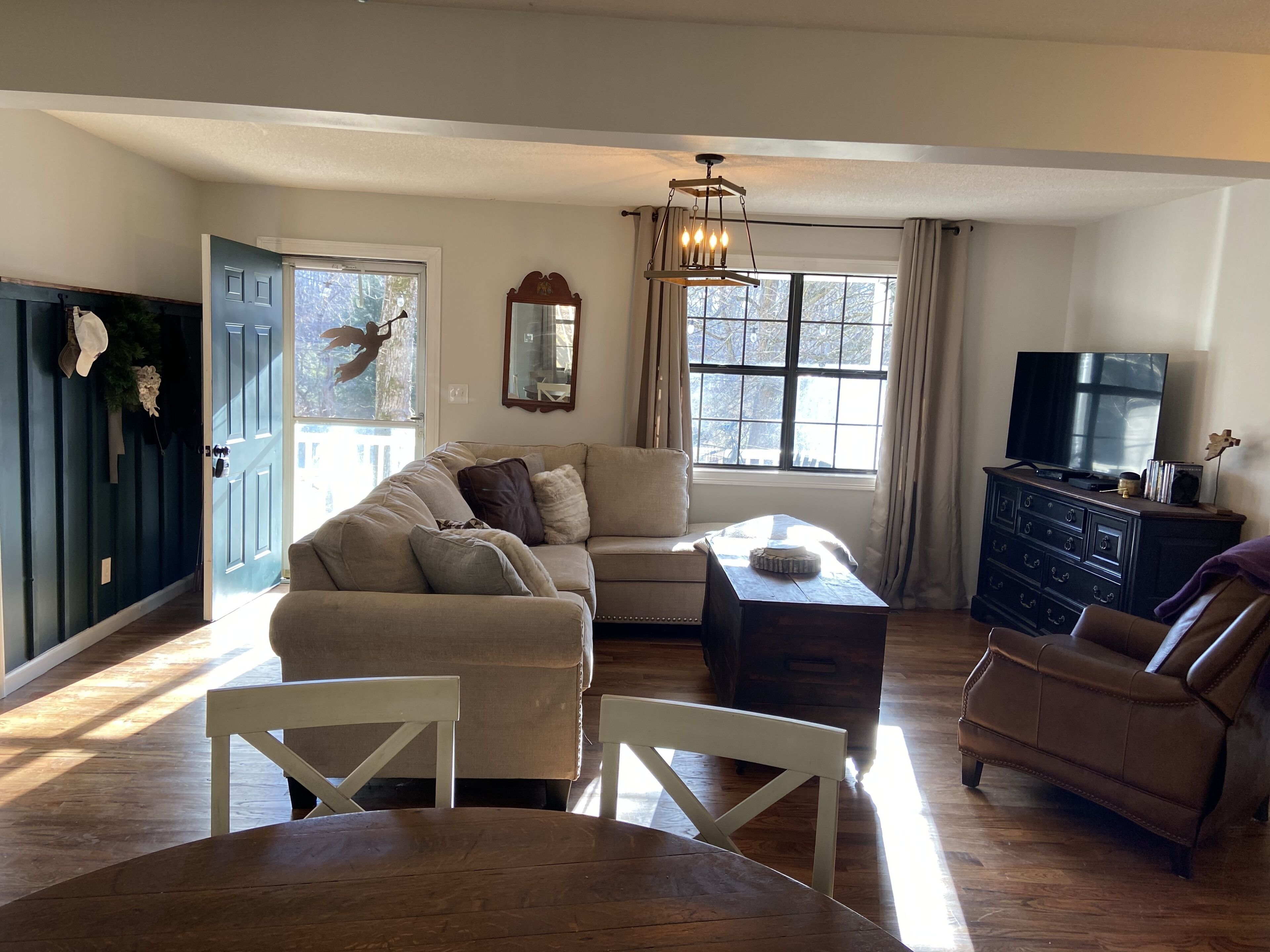 Un salon avec un canapé beige, une table basse en bois, un téléviseur posé sur une commode et une grande fenêtre avec des rideaux.