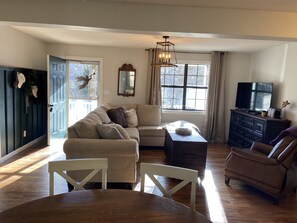 A living room with a beige sofa, a wooden coffee table, a TV on a dresser, and a large window with curtains.