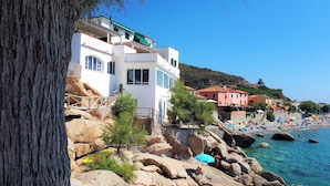 A coastal building with a balcony, a rocky shoreline, and a beach with umbrellas. - "SEA HOUSE" DIRECTLY ON THE SEA (Pomonte)