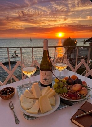 A table with a bottle of wine, glasses, cheese, fruits, and nuts, set against a sunset view.