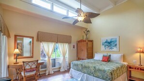 Un dormitorio con cama, ventilador de techo, ventana con cortinas, aparador de madera, una silla, mesa con lámpara y un cuadro en la pared.