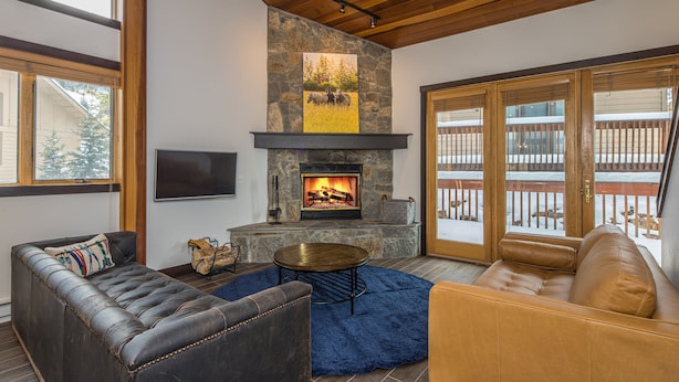A cozy living room with a stone fireplace, leather sofas, and a large window overlooking a snowy deck. - Ski In/Out home 50 steps to Bear Back Poma Lift w/steam shower - Big Horn Escape (Big Sky)