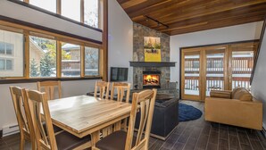 A modern living room with a wooden dining table, chairs, a fireplace, a sofa, and large windows. - Ski In/Out home 50 steps to Bear Back Poma Lift w/steam shower - Big Horn Escape (Big Sky)