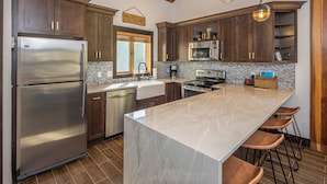 A modern kitchen with dark wooden cabinets, stainless steel appliances, a large marble island, and bar stools. - Ski In/Out home 50 steps to Bear Back Poma Lift w/steam shower - Big Horn Escape (Big Sky)