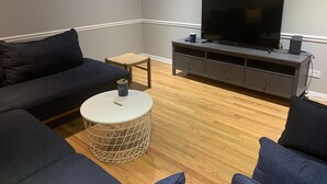 A living room with a dark blue sofa, a small round coffee table, a wicker side table, a grey entertainment unit with a flat-screen TV, and a wooden floor. - Home Sweet Home, minutes to Great Lakes Naval Base (Lake Bluff)