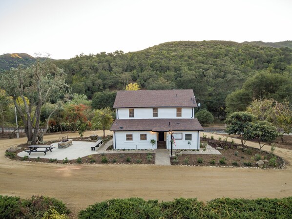 A two-story white house with a dark roof, surrounded by a landscaped garden with a picnic table and a small American flag.
