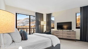Una camera d'albergo moderna con un letto grande, una TV a schermo piatto, un comò e vista su un paesaggio innevato attraverso le finestre.