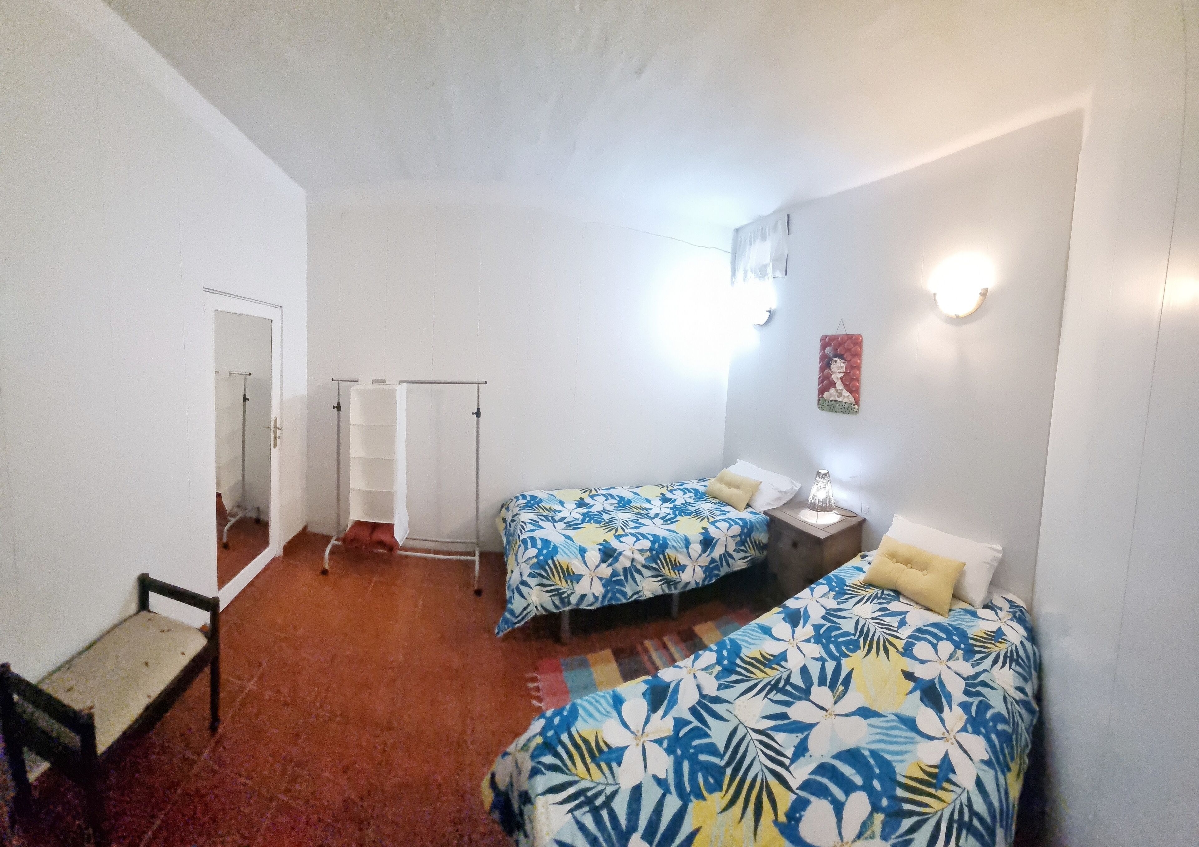 A room with two beds, a small table, a nightstand, a wall-mounted light, and a door leading to a bathroom.