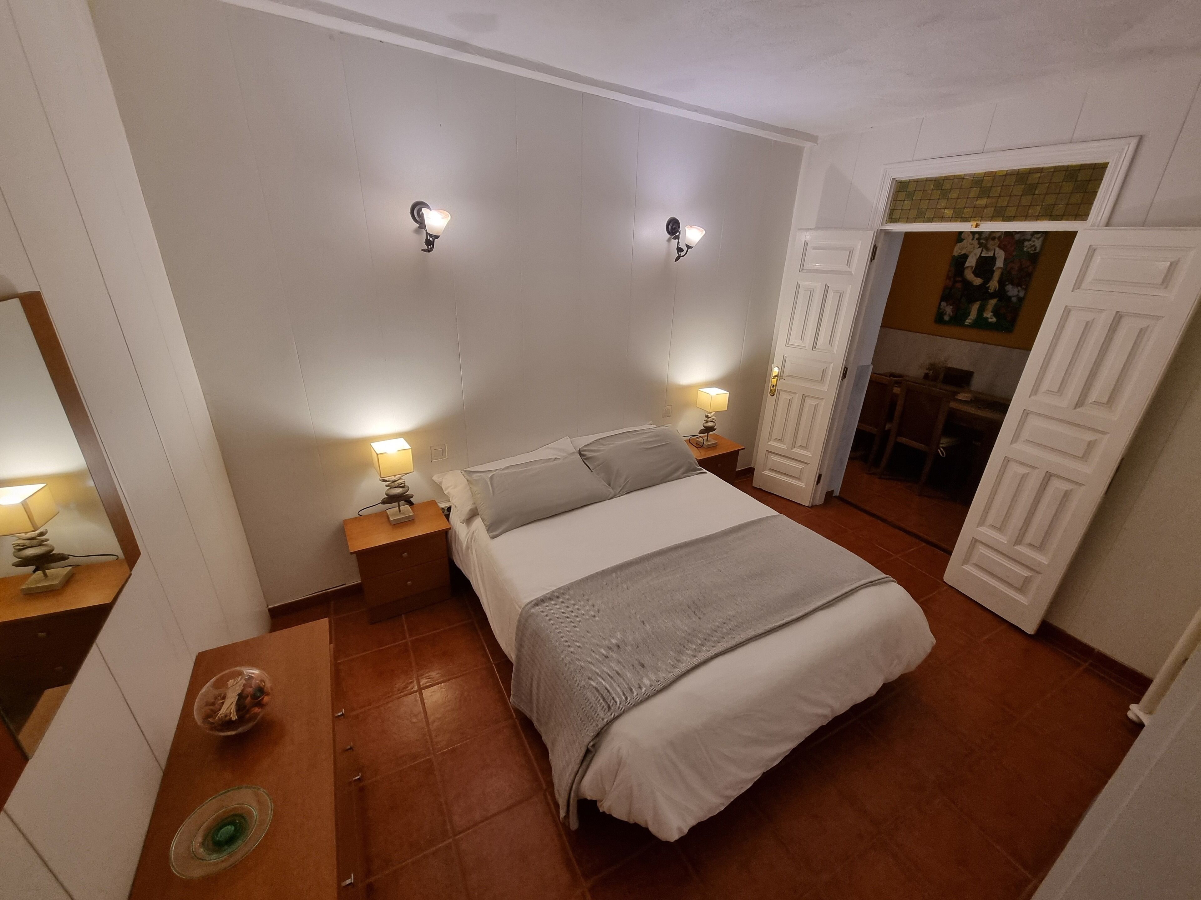 A bedroom with a bed, two bedside tables with lamps, a wooden nightstand, a small table with a bowl, and a door leading to another room.