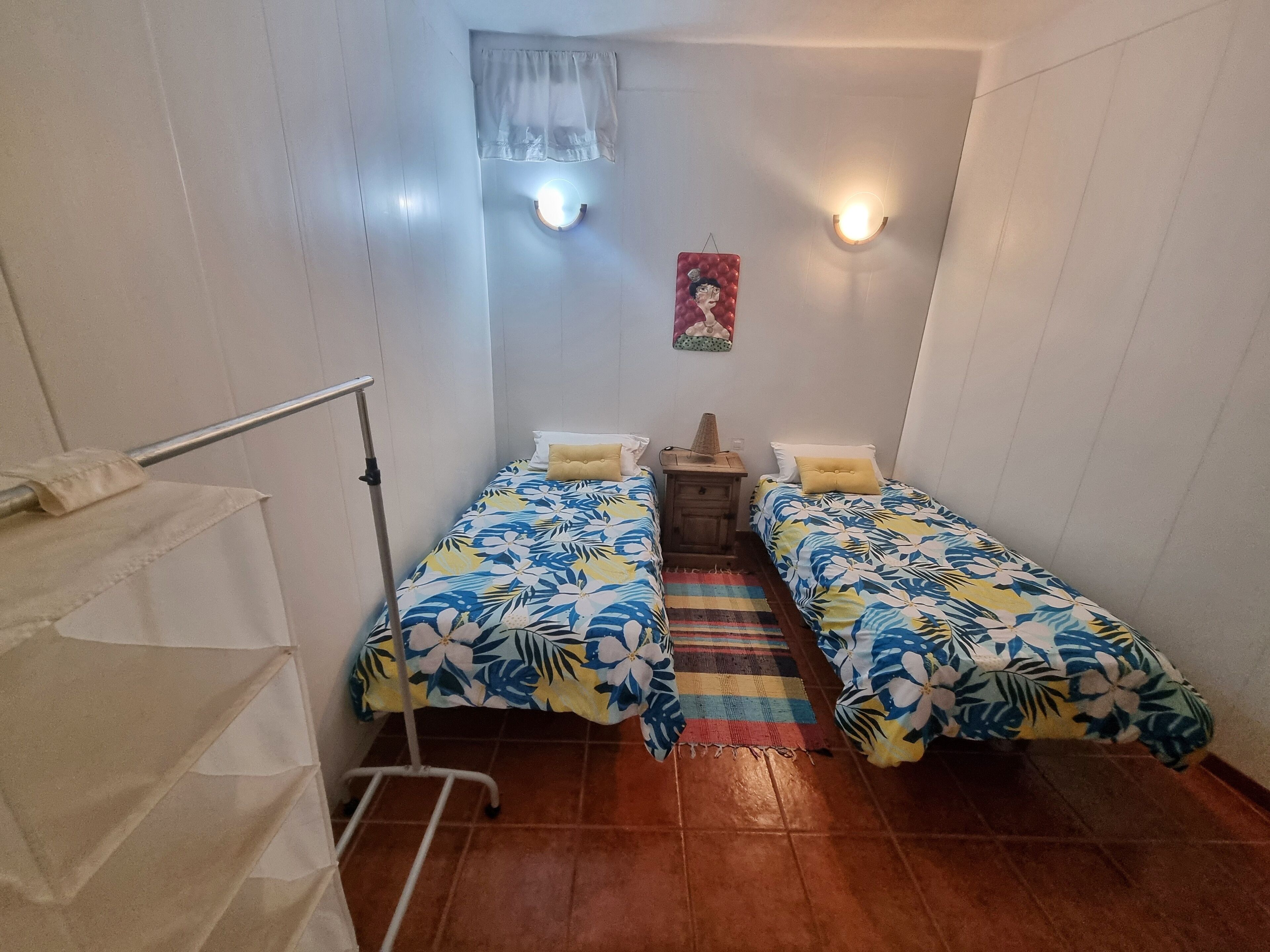 Une petite pièce bien éclairée, avec deux lits simples, une table de chevet en bois, un tapis rayé aux couleurs vives et un luminaire fixé au mur.