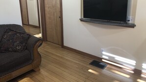 Een kamer met een houten vloer, een bank met een gestreept hoesje, een aan de muur bevestigde televisie en een deur met een houten omlijsting.