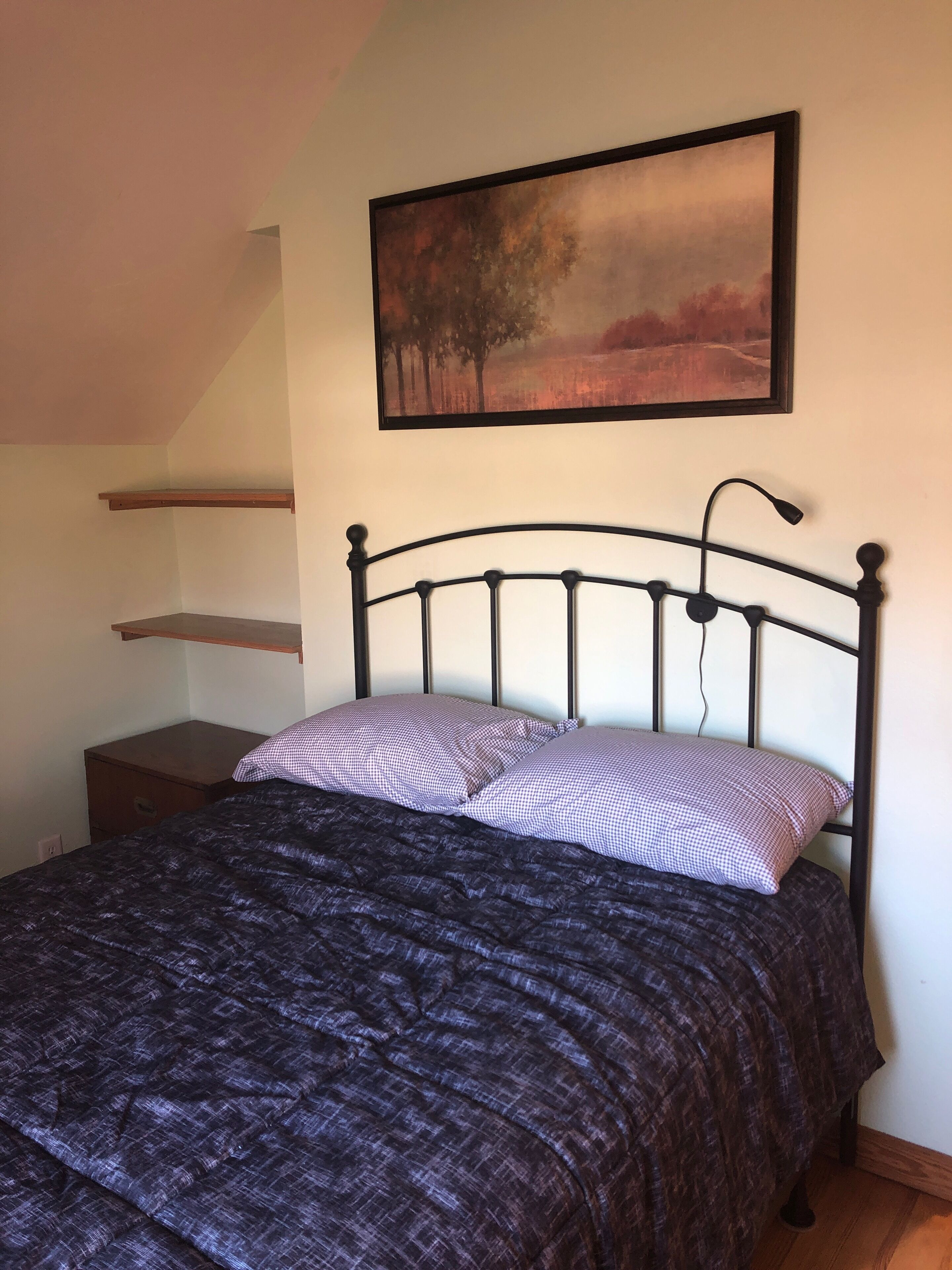 A bedroom with a bed, a nightstand, a shelf, and a painting on the wall.