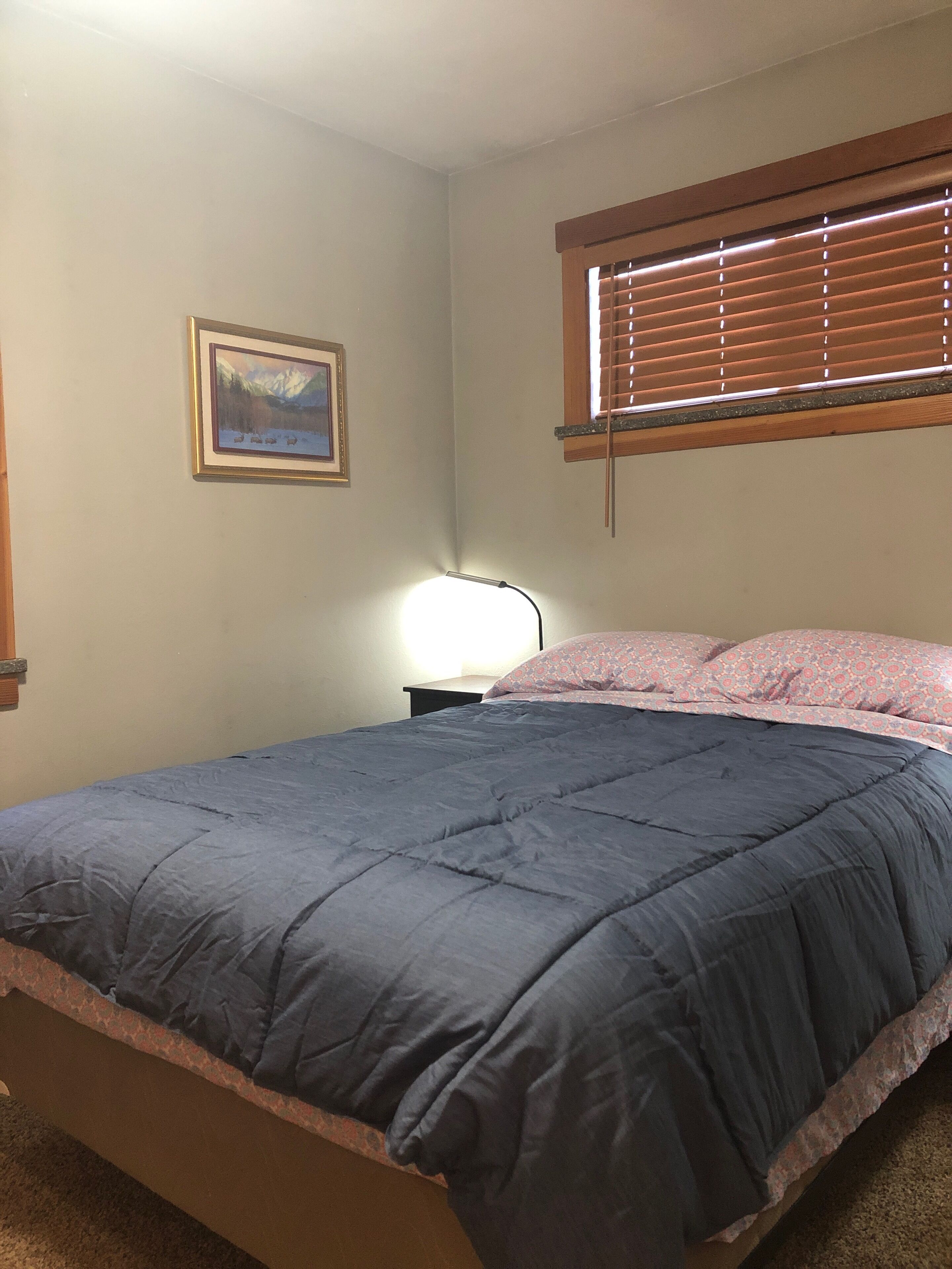 A bedroom with a bed, a nightstand, a lamp, a window with blinds, and a framed picture on the wall.
