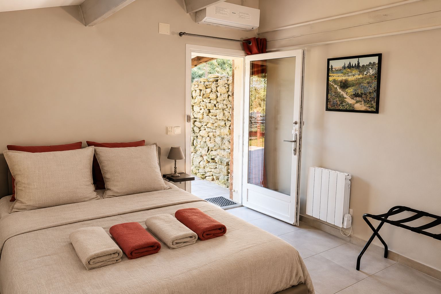 Un dormitorio con cama, mesita de noche, lámpara, radiador, puerta que da a un balcón y un cuadro en la pared.