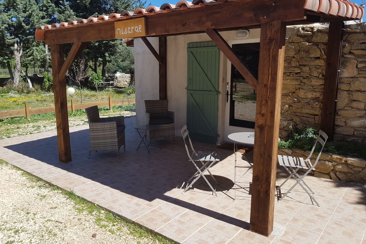 A wooden pergola with a sign reading "mistrea" in a garden area with outdoor seating and a stone wall.