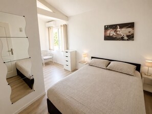 Un dormitorio con cama, una silla, cómoda, mesita de noche con lámpara y un cuadro en la pared.