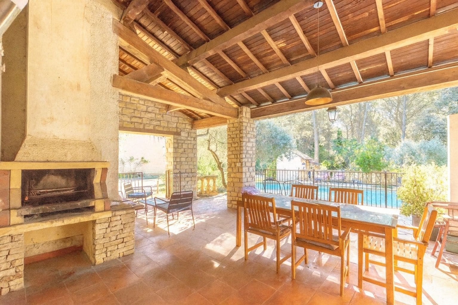 An outdoor patio with a brick fireplace, wooden furniture, and a view of a swimming pool and greenery.