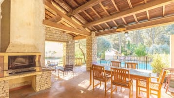 An outdoor patio with a brick fireplace, wooden furniture, and a view of a swimming pool and greenery.