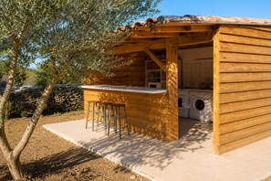 Cocina exterior de madera con barra y taburetes, lavadora y un muro de piedra al fondo.