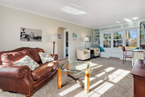 A living room with a brown leather sofa, a glass coffee table, and a white armchair. There is a bookshelf with various items and a window with a view of greenery.
