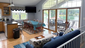A spacious living area with a dining table, chairs, a sofa, and a large window offering a view of a lake.