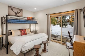 A room with a bunk bed, a single bed, a wooden table, and a view of a snowy landscape through a glass door.
