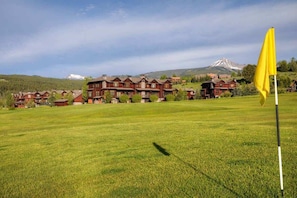 A golf course with a yellow flag, surrounded by lodge buildings and a mountain in the background.