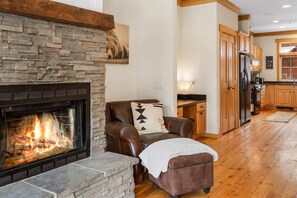 A living room with a stone fireplace, a leather armchair, and a kitchen in the background.