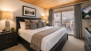 Un dormitorio con techo de madera, cama con ropa de cama blanca y almohadas estampadas, mesita de noche con lámpara, cuadro en la pared y balcón con mobiliario exterior.