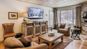 Una sala de estar acogedora con techo de madera, televisión, chimenea y un ventanal con vistas a un bosque nevado.