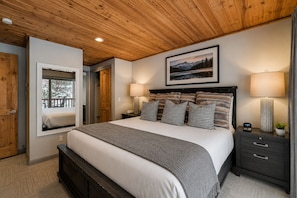 Un dormitorio con techo de madera, una cama con almohadones estampados, una lámpara de noche y un cuadro en la pared.