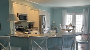 A modern kitchen with white cabinetry, a central island with bar stools, and a dining area with a table and chairs. - Beautiful spacious family friendly home in Rehoboth Beach with Pool (Rehoboth Beach)