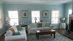 A living room with a white sofa set, a coffee table, and a patterned rug. There are three windows with blinds, a fireplace, and wall art. - Beautiful spacious family friendly home in Rehoboth Beach with Pool (Rehoboth Beach)