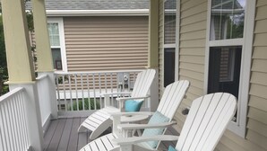 A covered porch with white Adirondack chairs and a small table, featuring a view of a house in the background. - Beautiful spacious family friendly home in Rehoboth Beach with Pool (Rehoboth Beach)