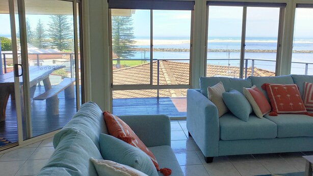 Ein Wohnzimmer mit einem hellblauen Sofa, einem Holztisch und Blick auf Strand und Meer durch große Fenster.