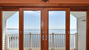 A wooden door with glass panels leading to a balcony with a white railing, overlooking a body of water. - Entire Waterfront Home with Panoramic Views of the Bay and Private Beach (Mastic Beach)