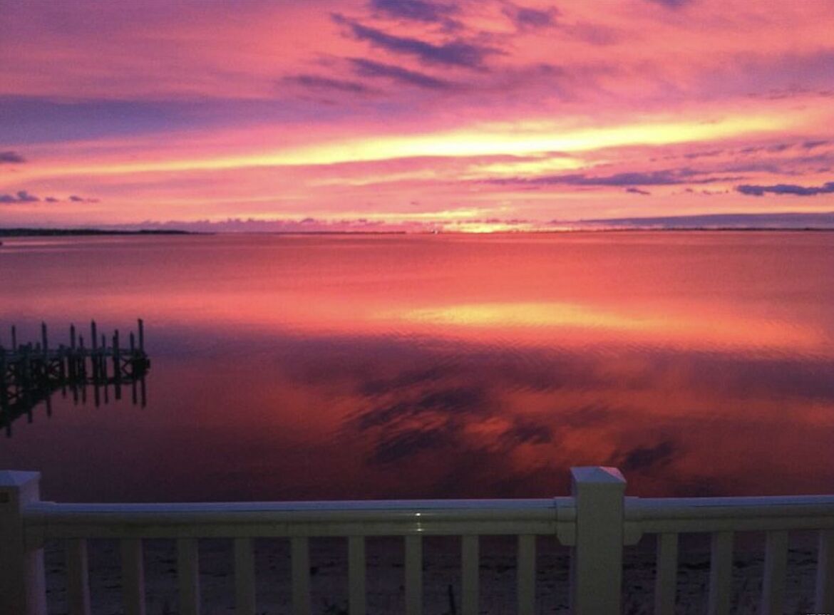 Un coucher de soleil sur une étendue d’eau calme, avec une jetée en bois au premier plan.