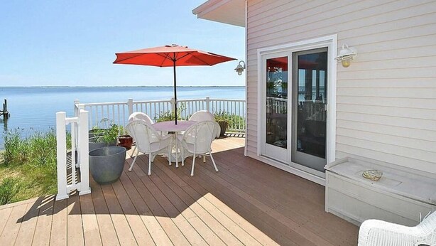 A wooden deck with a table and chairs, a red umbrella, and a view of the water. - Entire Waterfront Home with Panoramic Views of the Bay and Private Beach (Mastic Beach)