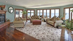 A spacious living room with a large window overlooking a body of water, a fireplace, and several pieces of furniture. - Entire Waterfront Home with Panoramic Views of the Bay and Private Beach (Mastic Beach)