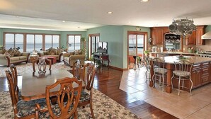 A spacious living room with a large dining table and chairs, a kitchen with a central island, and a view of the ocean through large windows. - Entire Waterfront Home with Panoramic Views of the Bay and Private Beach (Mastic Beach)