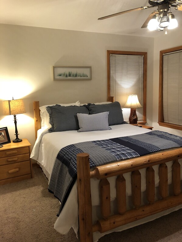 Een slaapkamer met een houten bed, twee nachtkastjes met lampen, een plafondventilator en een raam met jaloezieën.