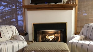 Ein Wohnzimmer mit Kamin, zwei gestreifte Sofas, einem Couchtisch mit Zeitschriften und einer tablettförmigen Ablage mit Sternenmuster sowie einem Fernseher, der über dem Kamin montiert ist.