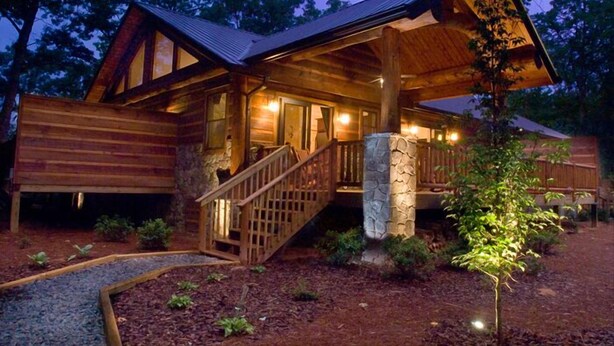 Une cabane rustique en rondins de bois, avec des fondations en pierre, une terrasse en bois et un escalier menant à l’entrée.
