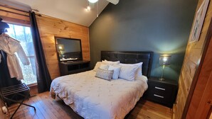 Une chambre avec un plancher en bois, un lit avec une literie blanche, un ventilateur de plafond, une table de chevet et un téléviseur fixé au mur.