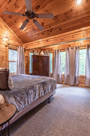 A wooden cabin bedroom with a bed, a ceiling fan, a wooden cabinet, and a window with curtains.