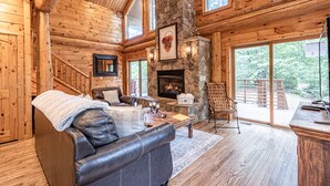 A log cabin interior with a stone fireplace, leather sofa, and large windows offering a view of the outdoors. - Watershed 1 - Modern-Rustic, Luxury, One King Bedroom Cabin (Bryson City)